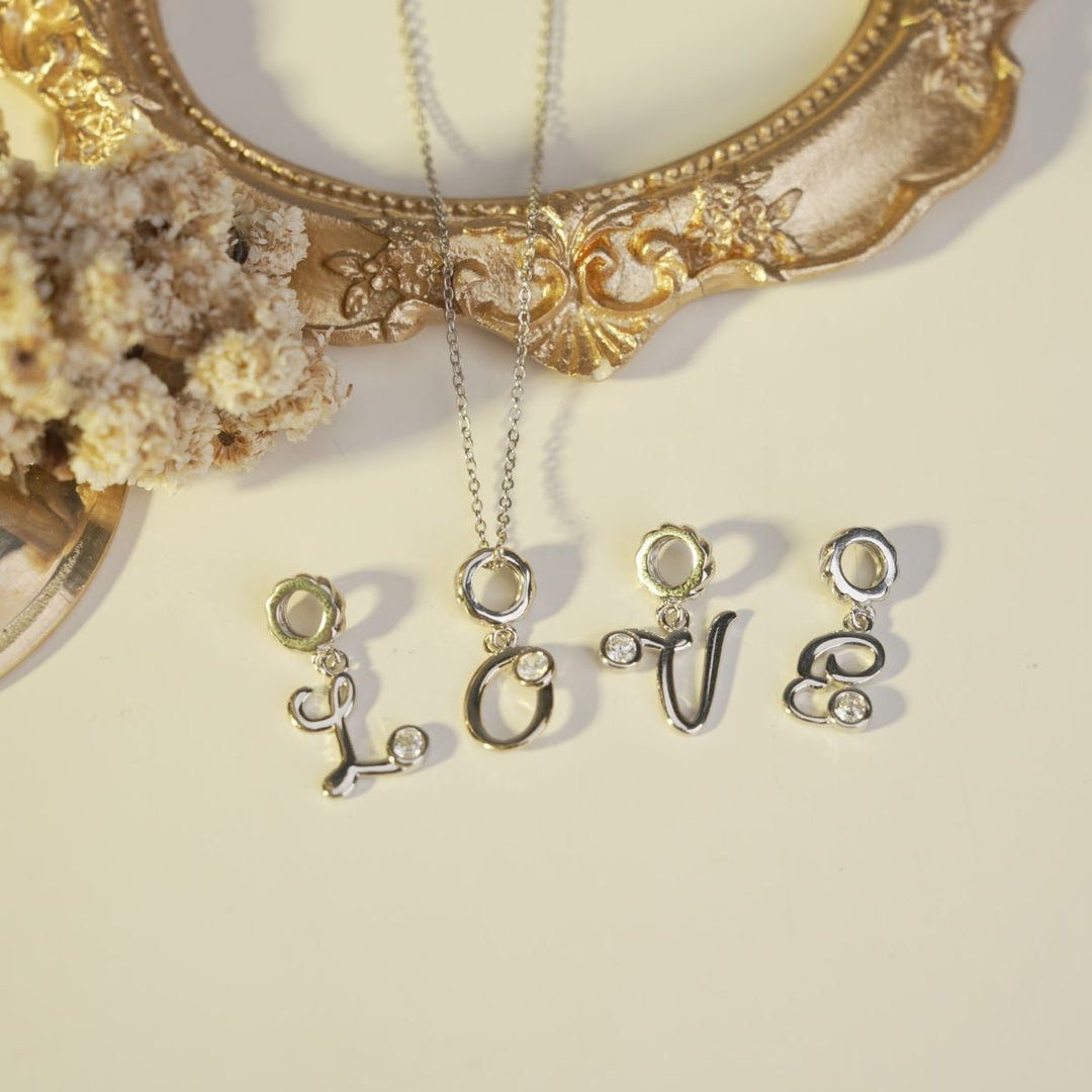 Liontin inisial bergaya kaligrafi membentuk kata LOVE berwarna silver dengan aksen zirconia, dipadukan dengan gantungan berbingkai bunga dan rantai perak, cocok sebagai hadiah romantis dan personal dari DAVINA.