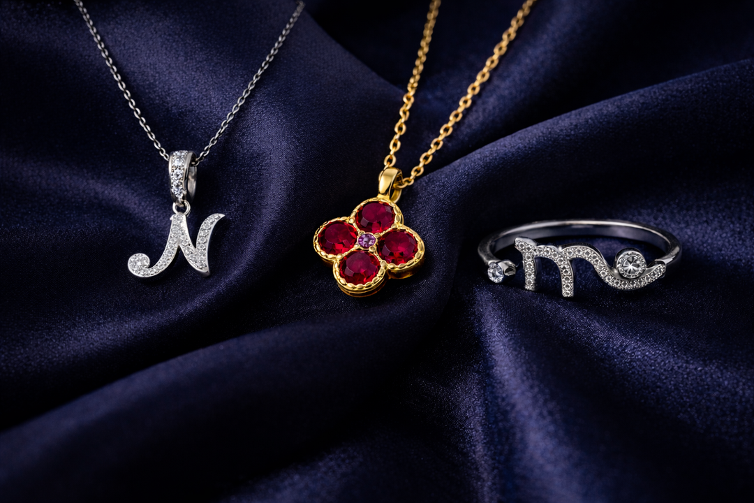 Perhiasan personal berupa kalung inisial huruf N, kalung birthstone merah, dan cincin zodiak berbahan perak, ditata elegan di atas kain biru gelap.