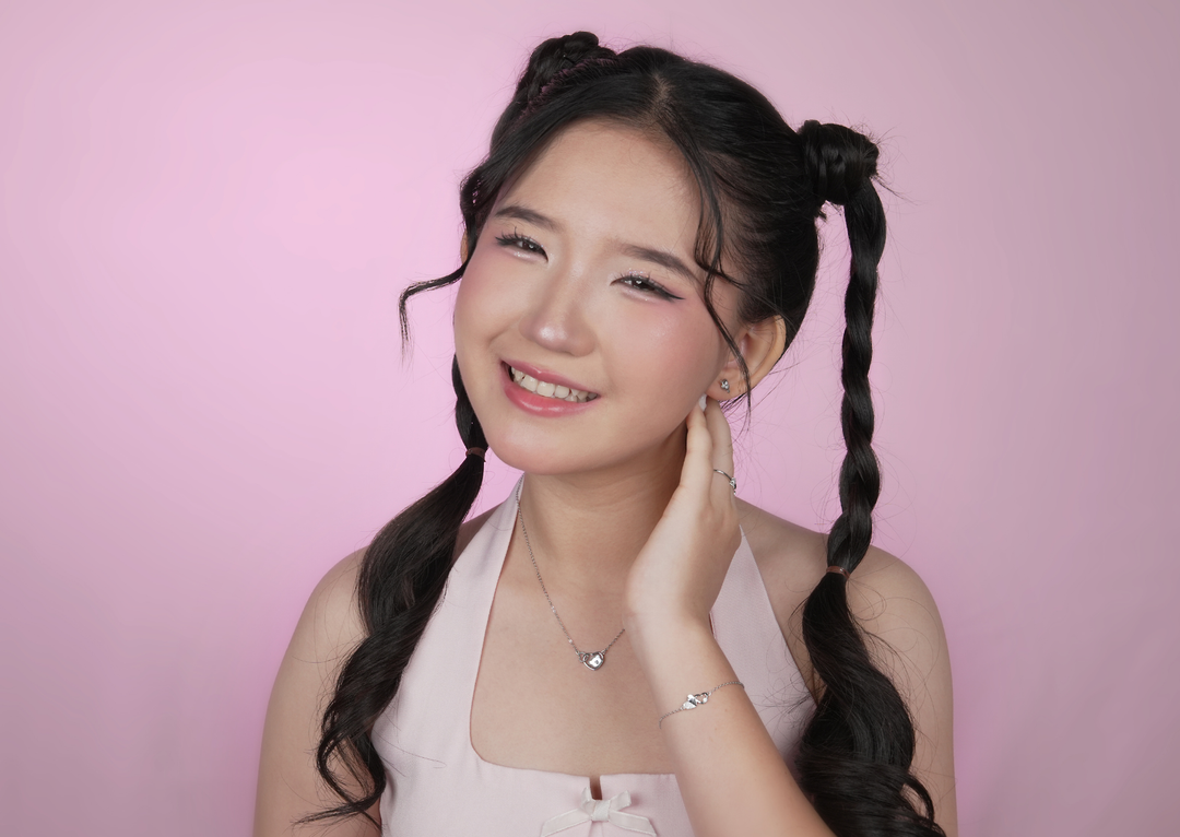 Model wanita dengan rambut kuncir dua memakai kalung liontin hati dan gelang perak S925 DAVINA, latar studio pink.