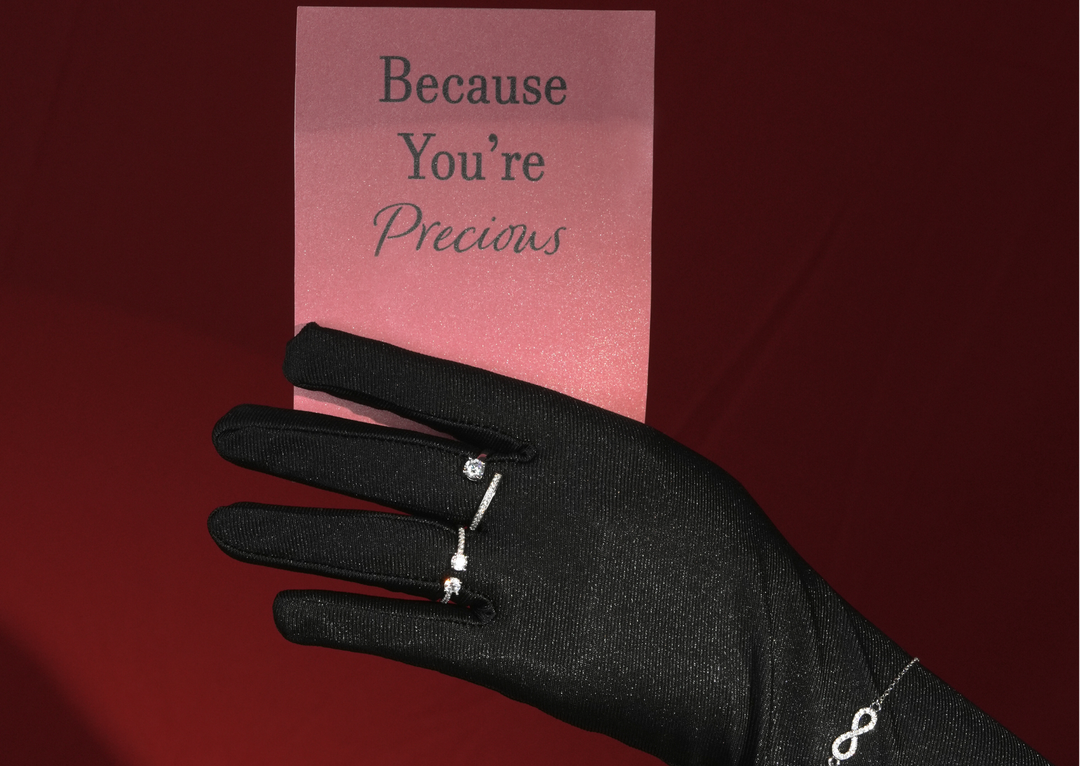 Tangan bersarung hitam memakai beberapa cincin silver 925 DAVINA sambil memegang kartu bertuliskan “Because You’re Precious” dengan latar merah.