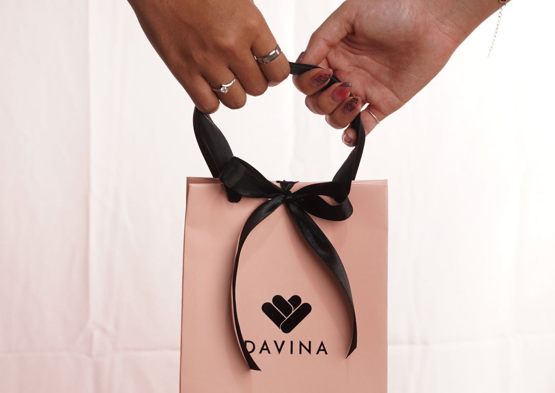Dua tangan memegang tas gift pink dari DAVINA dengan pita hitam dan logo DAVINA di depan