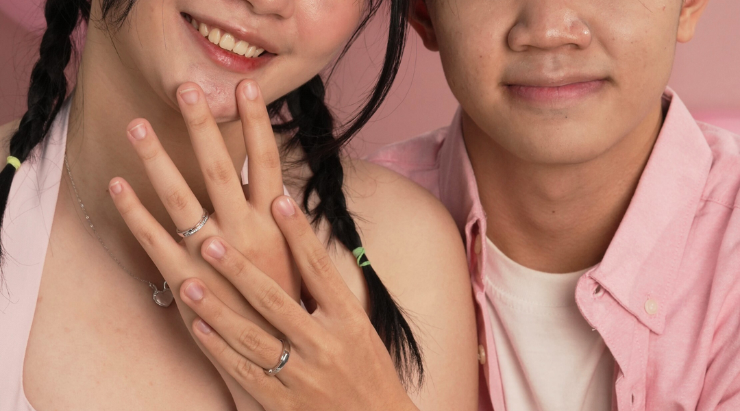 Pasangan memakai cincin couple perak S925, foto studio dengan latar pink, gaya pose romantis menampilkan cincin di jari.
